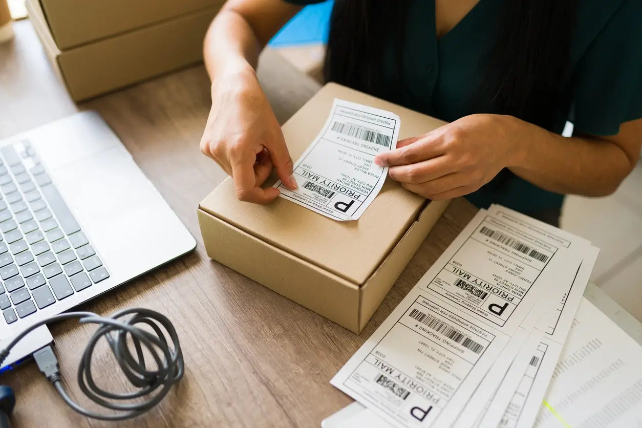 A person applies a standard shipping label to a cardboard box on a desk beside a laptop, with additional shipping labels nearby.